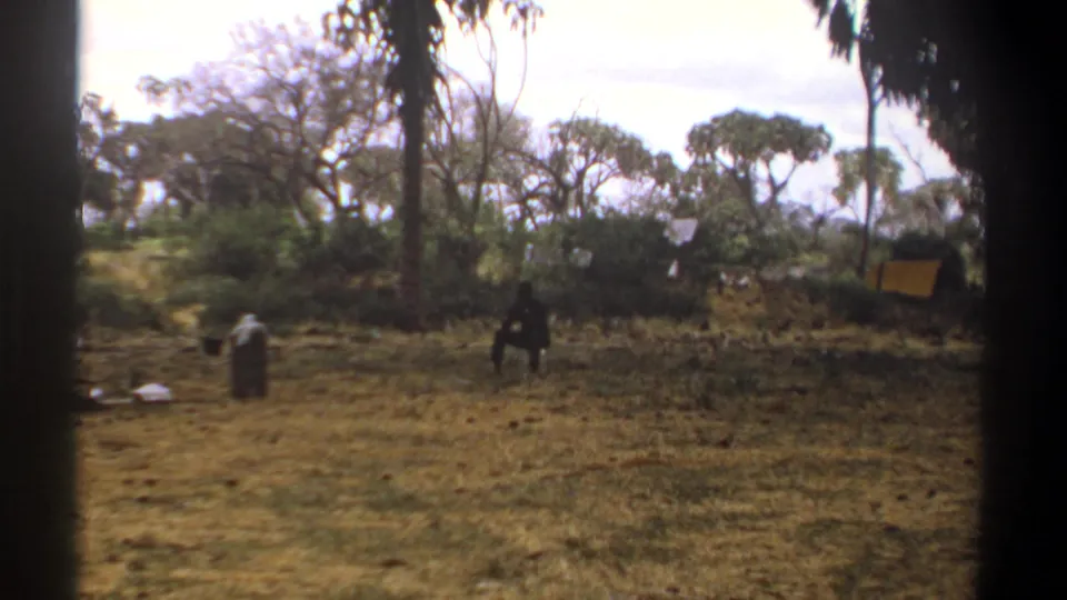 1969 Kenya frame showing distant figures in a dry clearing, with scattered containers, wooded background, and soft 8mm color.