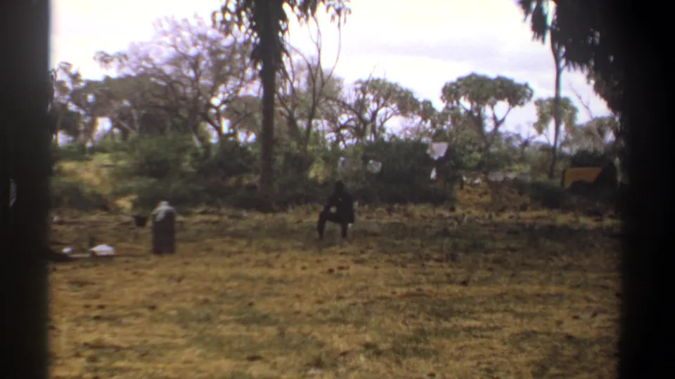 1969 Kenya 8mm frame showing a dry wooded campsite with a canvas tent, scattered gear, and a distant person in the clearing.