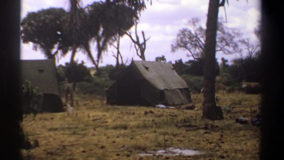 1969 Kenya 8mm frame showing two pitched tents in a dry woodland camp, with gear on the ground and trees beyond.