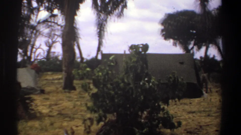 1969 Kenya 8mm frame showing a distant person in light clothing near canvas tents and trees at a campsite.