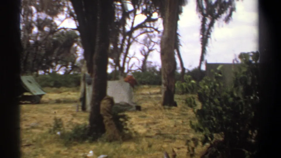 1969 Kenya home-movie frame with a distant person, canvas tents, dry grass, trees, and cloudy sky.