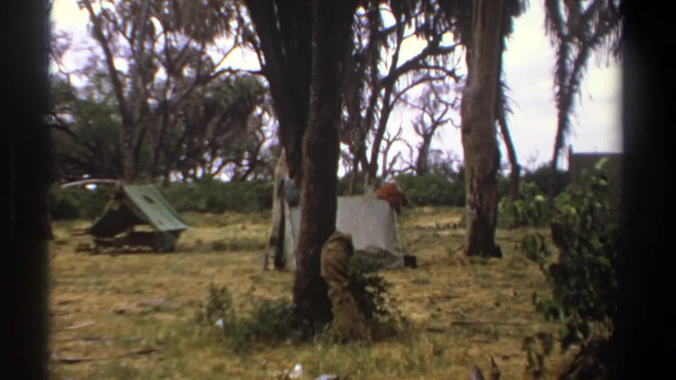 1969 Kenya campsite view with simple tents, tall trees, dry grass, and dark foreground edges from the 8mm frame.