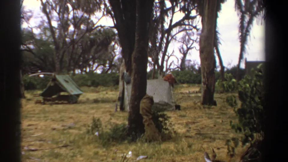 1969 Kenya home-movie frame of a rustic campsite, with two tents, tall trees, shrubs, and dry grass.