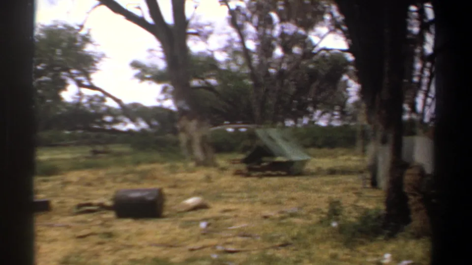 1969 Kenya 8mm frame with slight motion blur, showing a green tent, trees, dry grass, and scattered camp items.