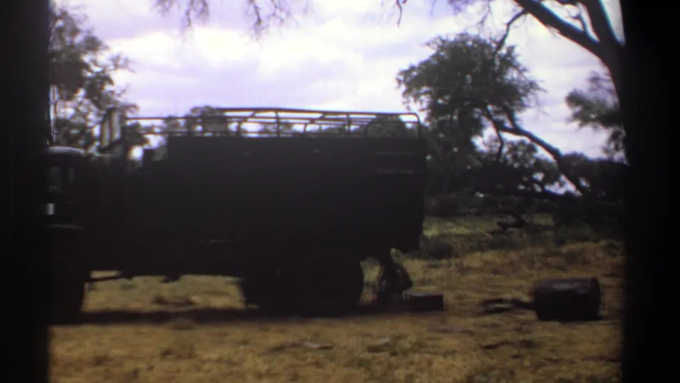 1969 Kenya 8mm frame with a large truck in profile and a person crouched near its underside at a wooded campsite.