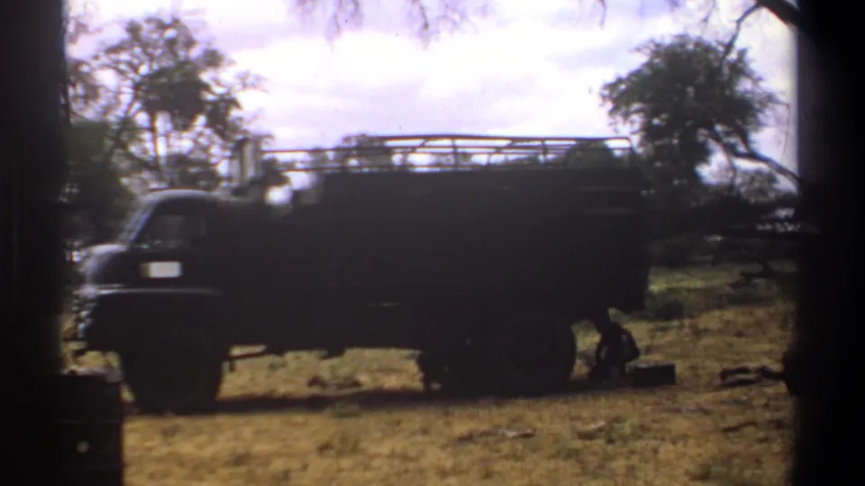 1969 Kenya 8mm frame showing the side of a large truck while a person works low near the vehicle in a dry camp area.