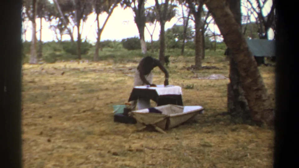 1969 Kenya 8mm frame of a person setting up or arranging a camp table beside folding canvas gear under trees.