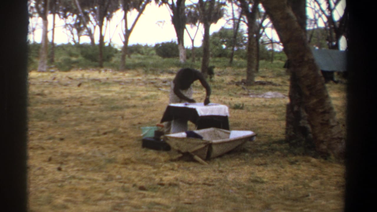In 1969 Kenya home-movie footage, a person leans over a small white-covered camp table in a wooded clearing.