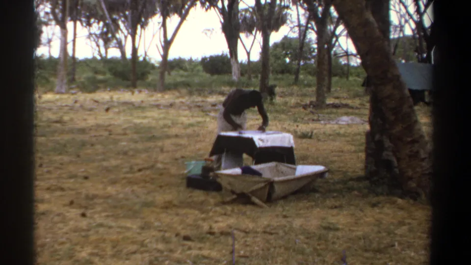 A person continues arranging items at a small covered table in a wooded Kenya camp setting, filmed on 8mm in 1969.