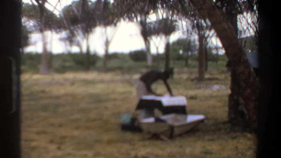 The 8mm frame falls soft as the person bends over the covered table in the Kenya camp clearing.