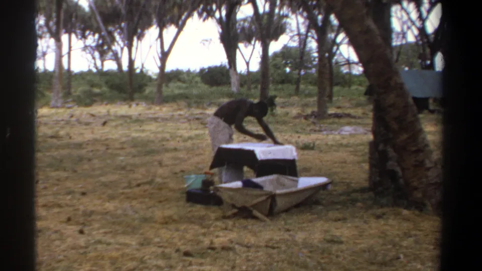 In the wooded camp clearing, the person reaches across the cloth-covered table while a low boat-like or canvas object sits nearby.