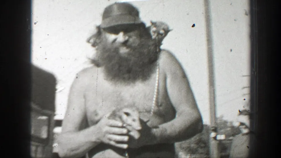 In 1939 California home-movie footage, a shirtless bearded man in a cap holds a small mouse-like animal while standing outdoors.