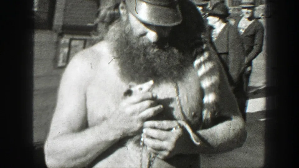 The man lowers his gaze toward the small animal in his hands, with street buildings and a bystander partly visible behind him.