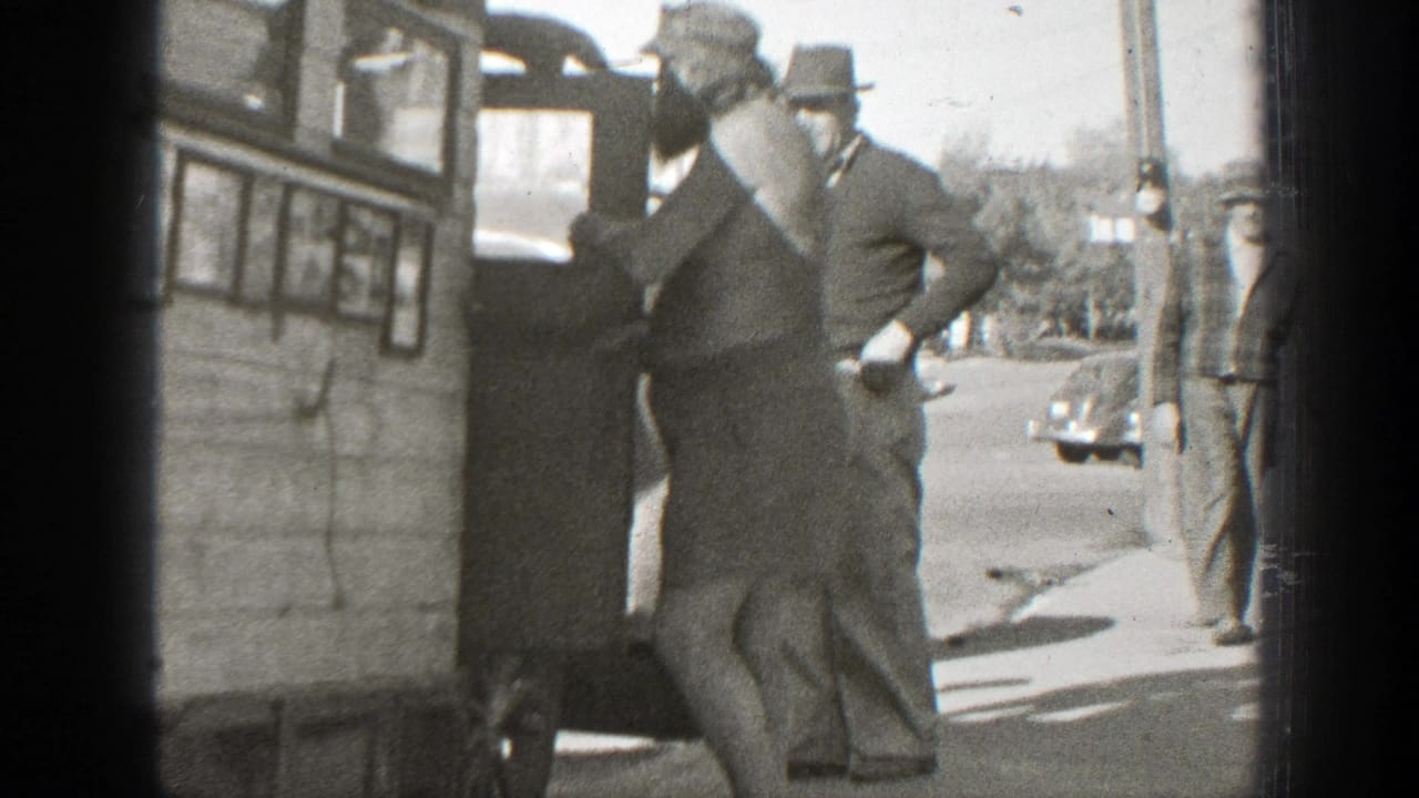 In 1939 California home-movie footage, adult men in hats watch as a shirtless man handles a small dog near a wooden vehicle or structure.