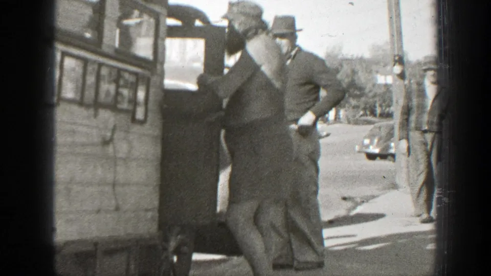 In 1939 California home-movie footage, adult men in hats watch as a shirtless man handles a small dog near a wooden vehicle or structure.