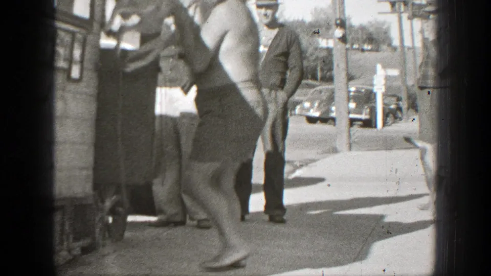 A shirtless man in shorts lifts a small dog near a wooden structure as several adult men watch on a California street corner.