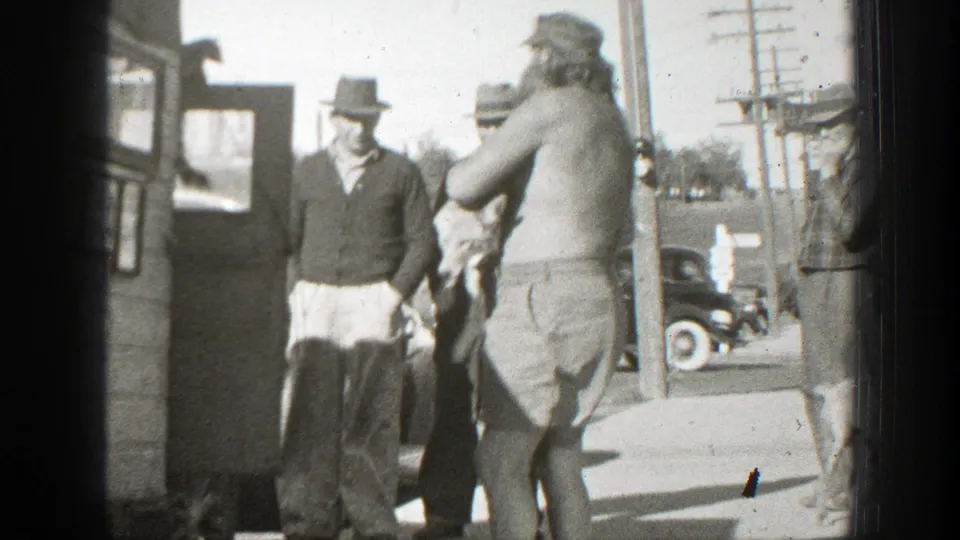 The shirtless man turns with the small dog in his arms while men in hats remain beside the wooden structure and vintage cars in the background.