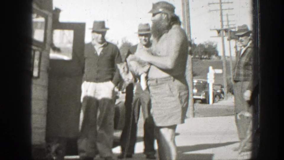 In the final frame, a shirtless man holds the small dog while men in hats stand near a wooden structure, parked car, and utility poles.
