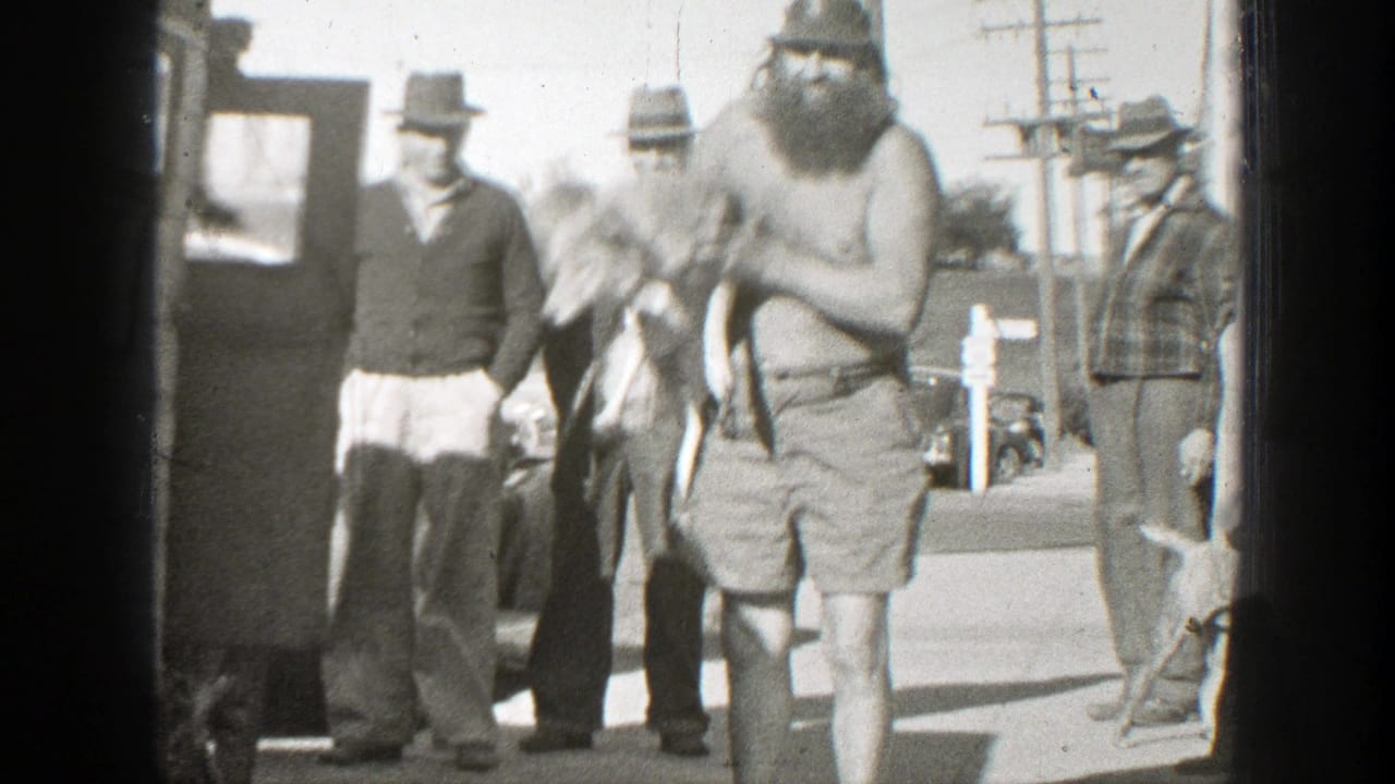 In 1939 California footage, a shirtless bearded man in shorts holds a small dog while several men in hats watch beside parked cars.