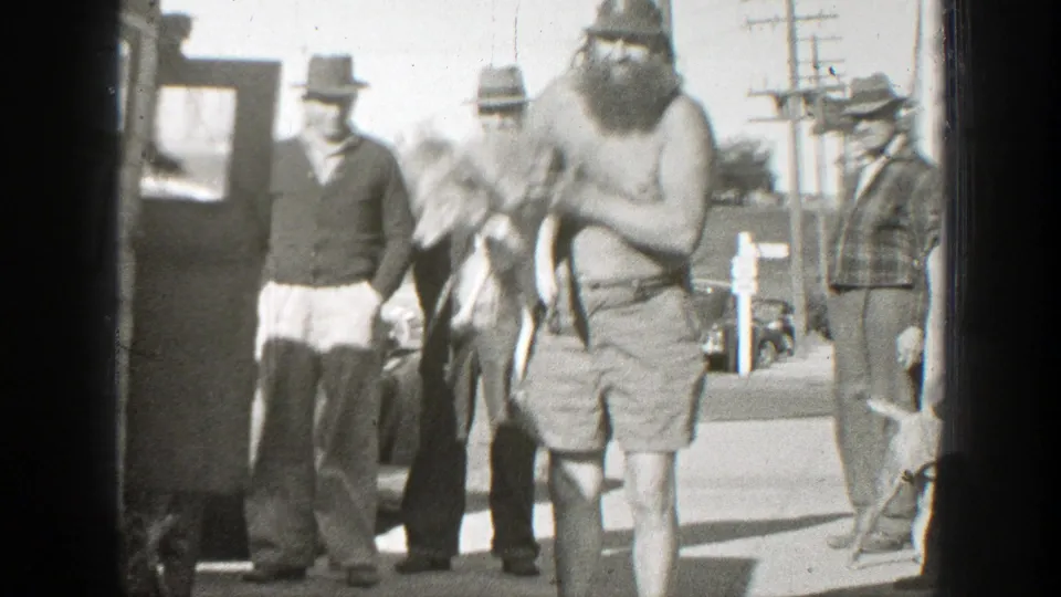 In 1939 California footage, a shirtless bearded man in shorts holds a small dog while several men in hats watch beside parked cars.