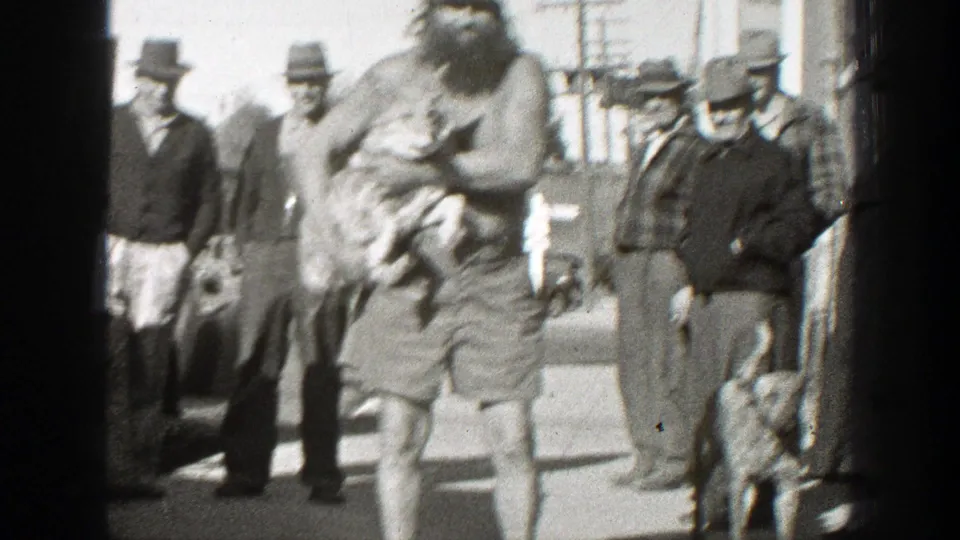 The bearded man moves across the frame holding a small dog, with several adult men in fedoras and street clothes visible behind him.