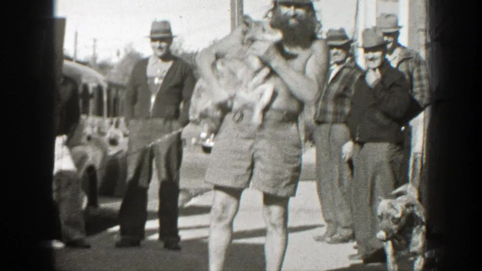 A shirtless bearded man cuddles a small dog near his face while men watch and another dog stands at the lower right of the street scene.