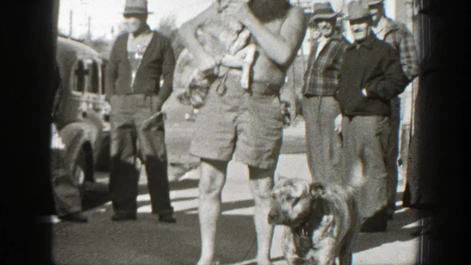 In the final frame, the man holds the small dog at his chest while another dog stands in front and several men look on from the sidewalk.