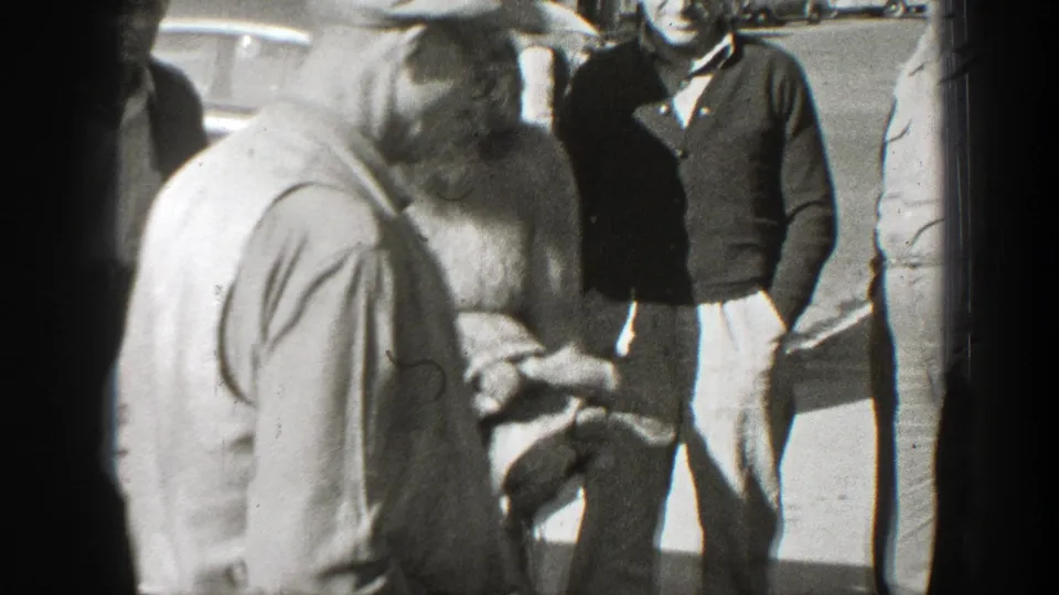 The bearded man looks down at a turtle in his hands, with several adult onlookers visible in the sunny California setting.