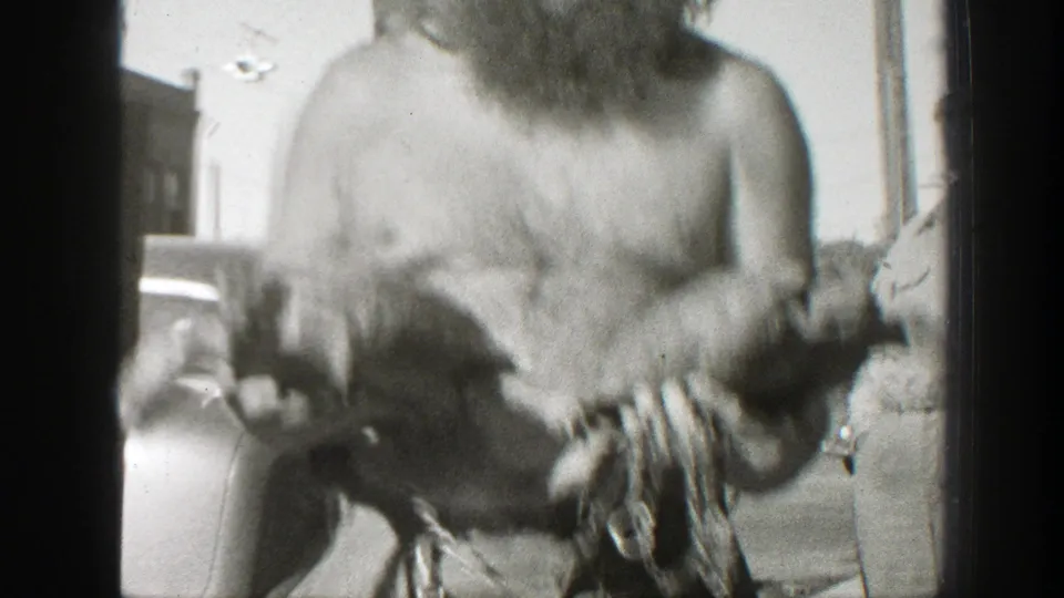 A closer 8mm frame shows the shirtless bearded man holding two turtles, with a vintage car and a standing adult partly visible behind him.