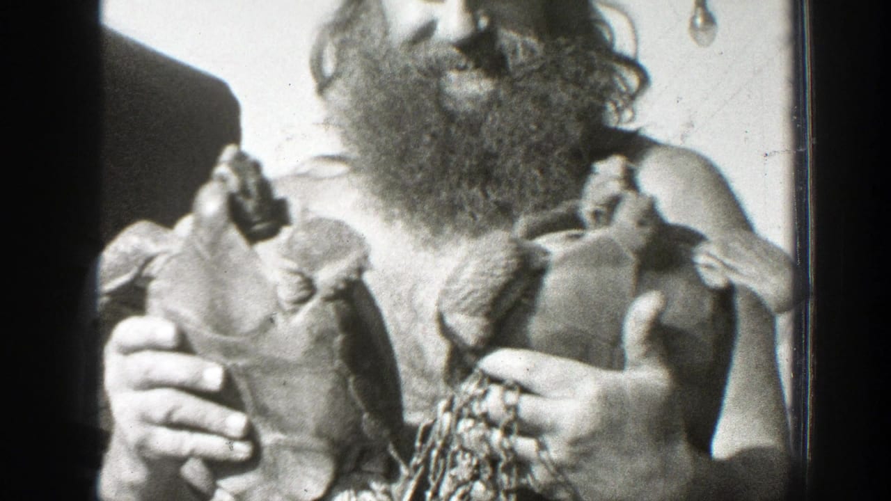 Bearded shirtless man holds two large turtles toward the camera.