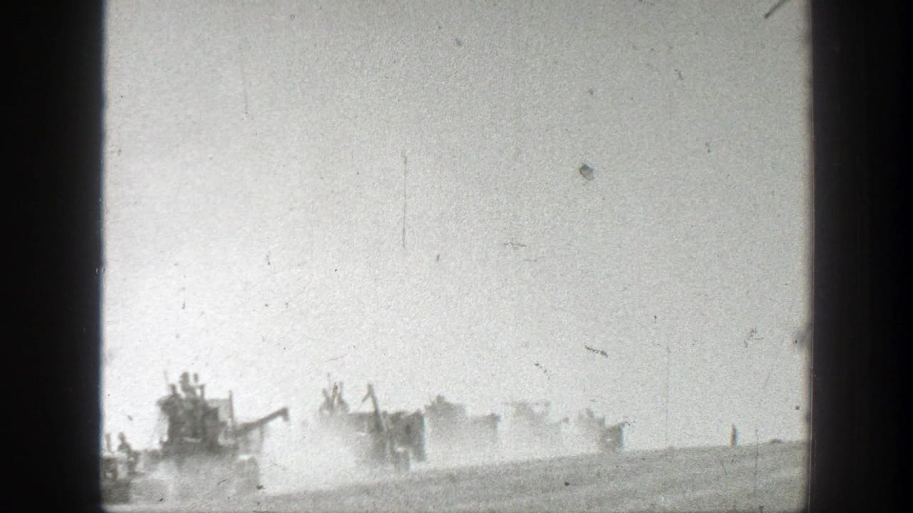 Distant harvest machines moving across a dusty field under a pale sky.