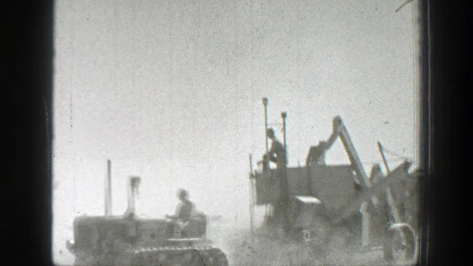 A tractor driver moves near the side of a large harvesting machine, with workers partly visible on the platform in 1939 California footage.