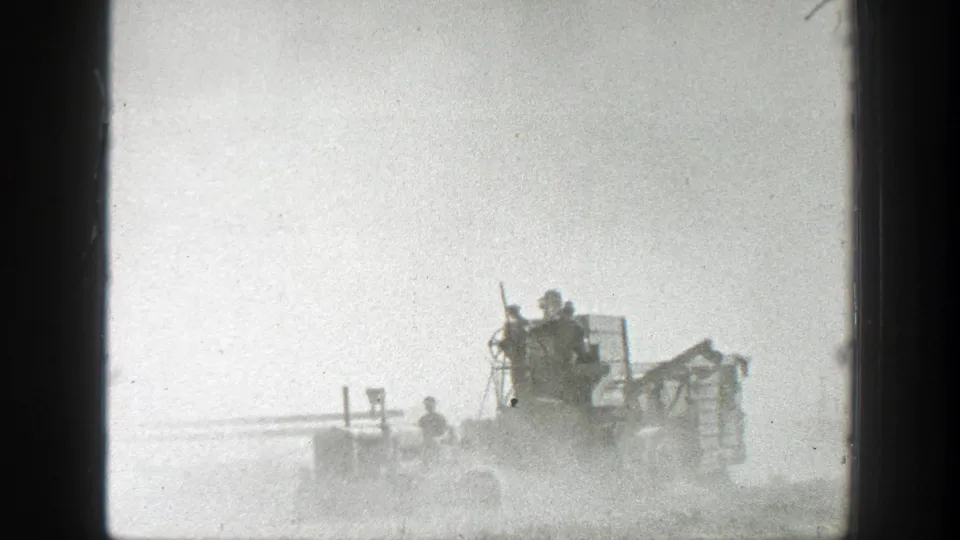 Old farming equipment crosses a dusty California field, with several people silhouetted on or near the machine.