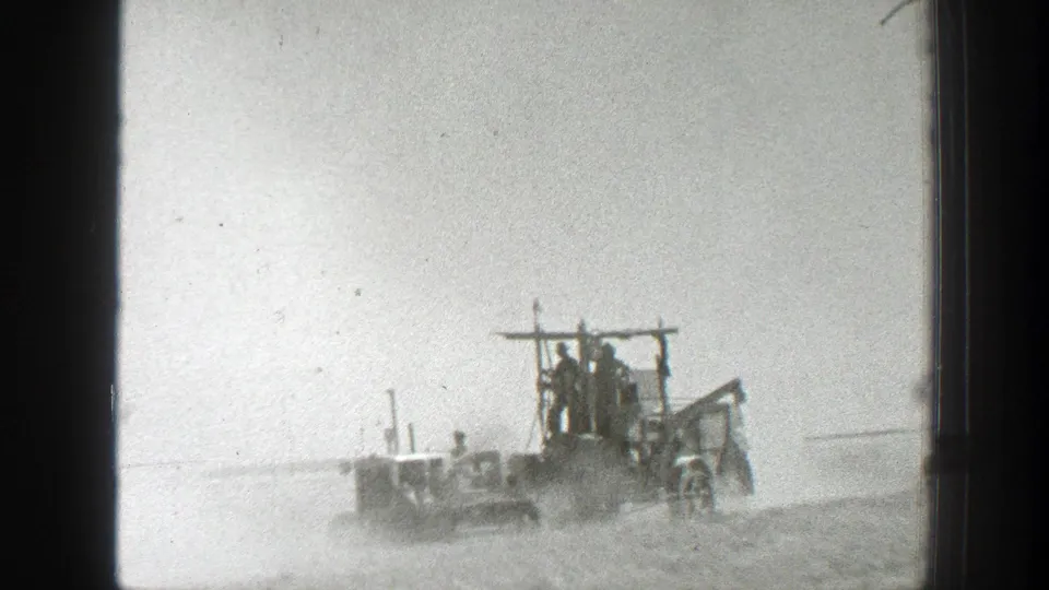 Black-and-white 8mm view of early farm equipment carrying several people across a dusty California field.