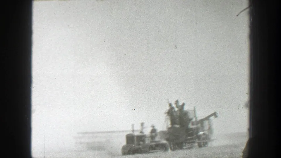 A distant 1939 California home-movie frame shows workers aboard a harvester moving through haze and dust in an open field.