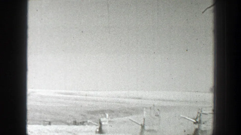 Wide 8mm home-movie frame of a bright agricultural field with parts of farm machinery at the bottom edge.