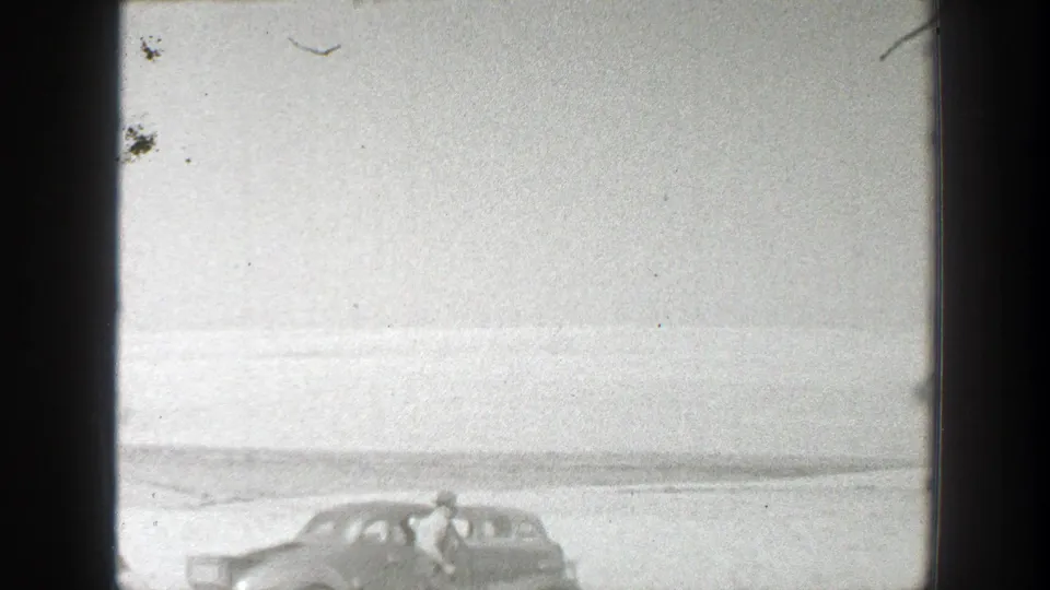 A rounded late-1930s style car appears in an open California field, with one person beside it in soft black-and-white footage.