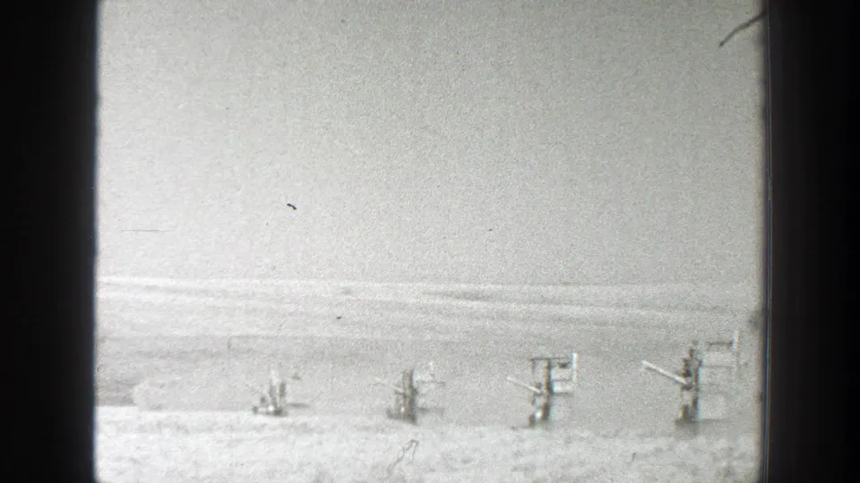 Grainy black-and-white frame of farm equipment working in a broad California field, shot from a distant fixed viewpoint.