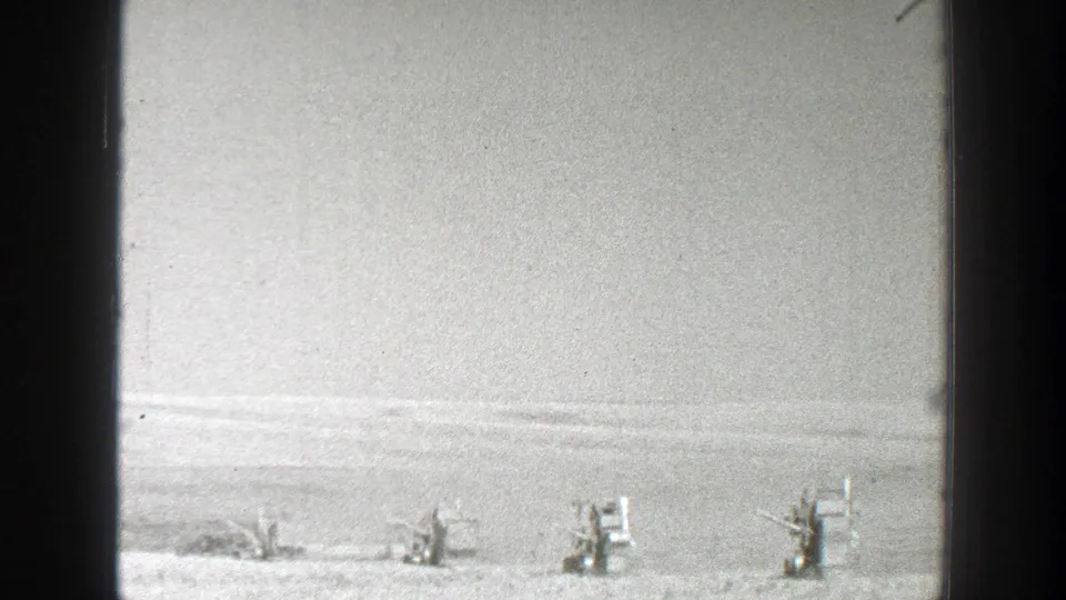 1939 California 8mm frame showing distant equipment or vehicles on an open plain with low hills faintly visible beyond.