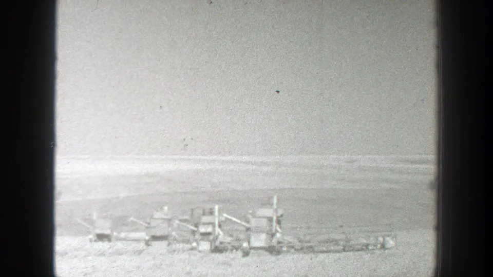 Black-and-white 8mm view of a broad California plain with a small line of equipment or vehicles near the horizon.