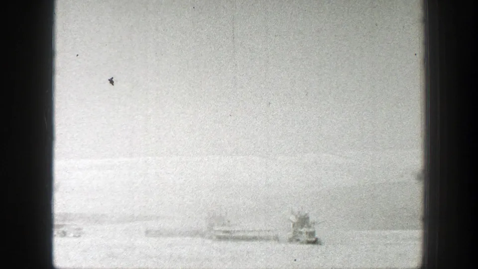1939 California 8mm frame with distant vessels or waterfront structures, pale water, low hills, and a bright hazy sky.