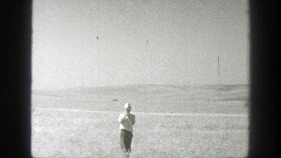 1939 California 8mm frame of a man in a white hat standing in open grassland, appearing to hold a camera or object near his face.