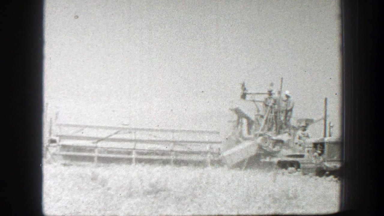 1939 California 8mm frame showing harvest machinery in a wheat field with several workers riding on elevated equipment platforms.