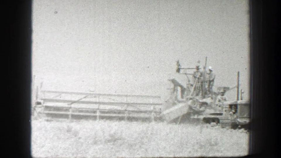 1939 California 8mm frame showing harvest machinery in a wheat field with several workers riding on elevated equipment platforms.