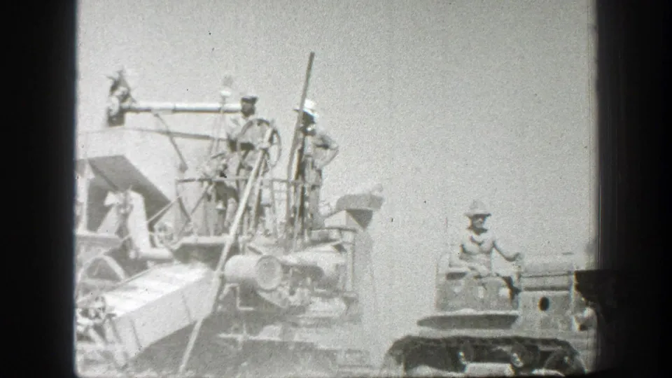 A tracked tractor or bulldozer pulls a combine across a California wheat field, with workers standing on the machinery in the 1939 footage.