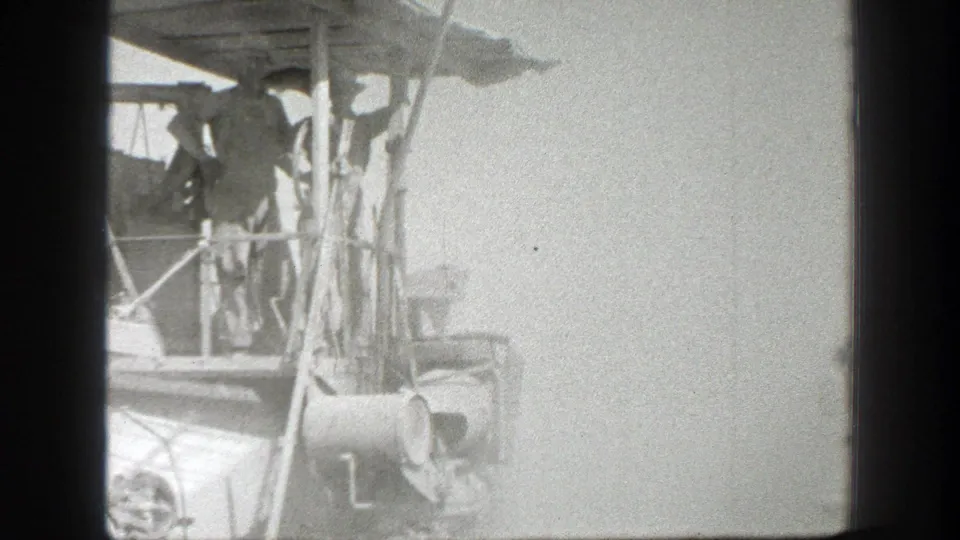Black-and-white 8mm frame from California 1939 showing workers near an elevated machine platform with posts, pipes, and railing.