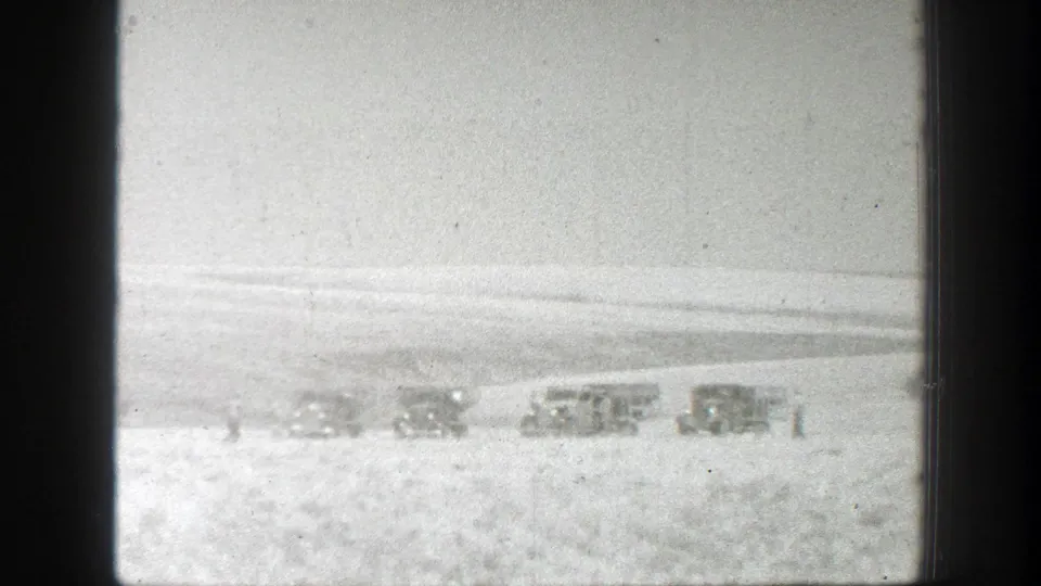 Black-and-white 8mm view of a California farm field with several distant machines or vehicles near the horizon.