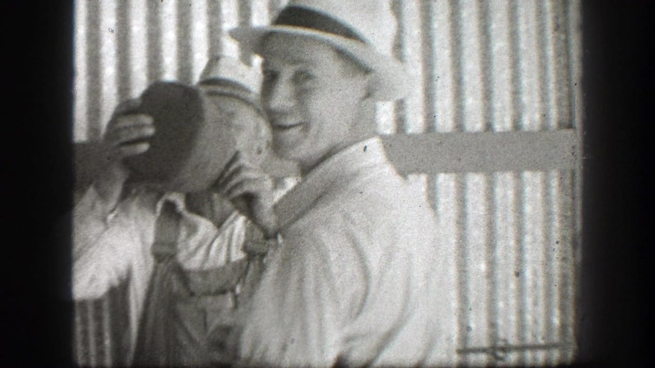 Two men in hats stand by corrugated metal as one drinks from a large jug.