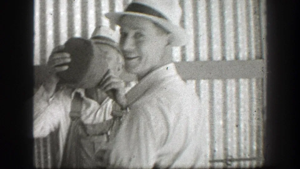 1939 California home-movie frame of two adult men in hats outdoors, one raising a large jug while the other smiles near a corrugated wall.