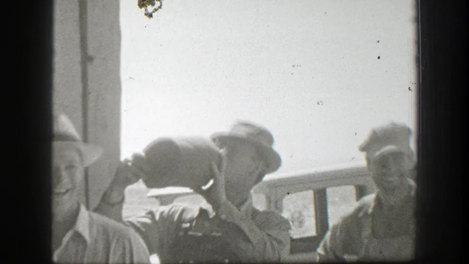 Overexposed 1939 California 8mm frame with several men in hats near a porch railing as one man tips a large jug upward.